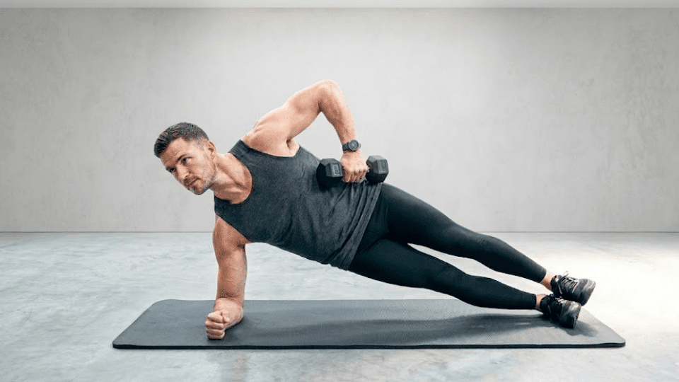 a muscular man performing weighted side plank with dumbbell on black yoga mat in minimalist studio