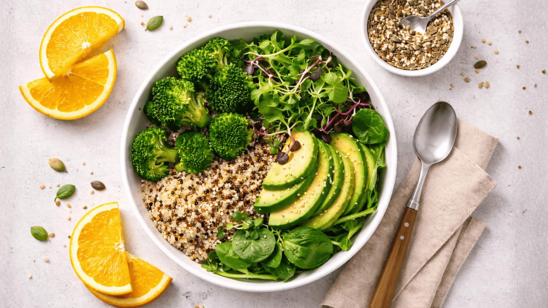 healthy bowl with broccoli, avocado, quinoa, greens, orange slices, seeds, and spoon on light grey surface
