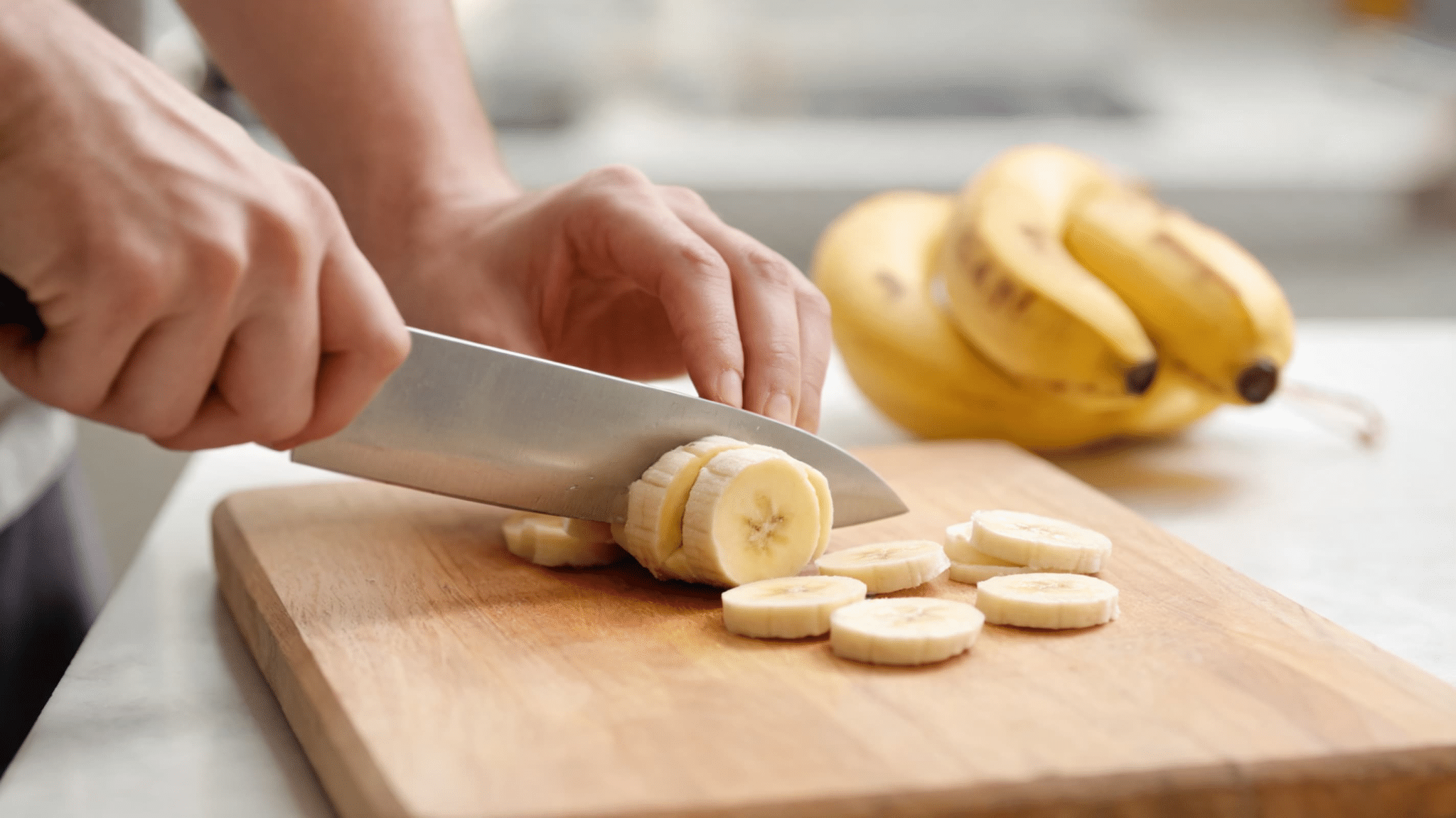 A person slices a banana on a wooden cutting board in a bright kitchen. The focus is on the knife and banana,