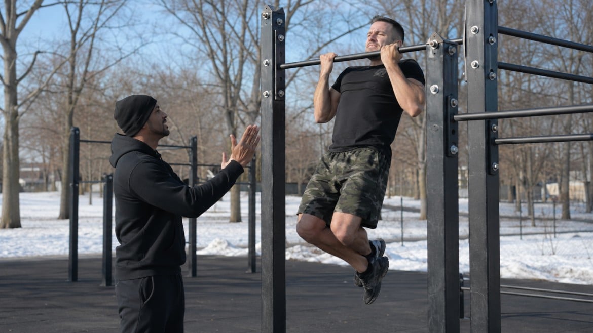 A man in camouflage shorts does a pull-up on outdoor bars, while a trainer in black clothes encourages him