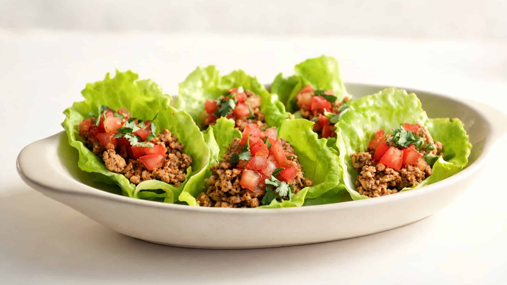 A white oval plate holds lettuce leaf wraps filled with seasoned ground meat, topped with diced tomatoes and cilantro