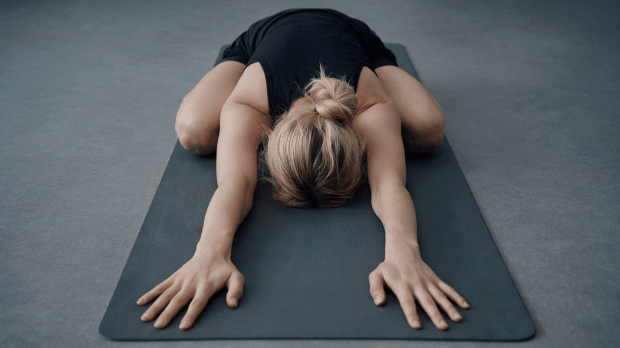 A woman in a black outfit performs a yoga child's pose on a gray mat, arms extended forward.