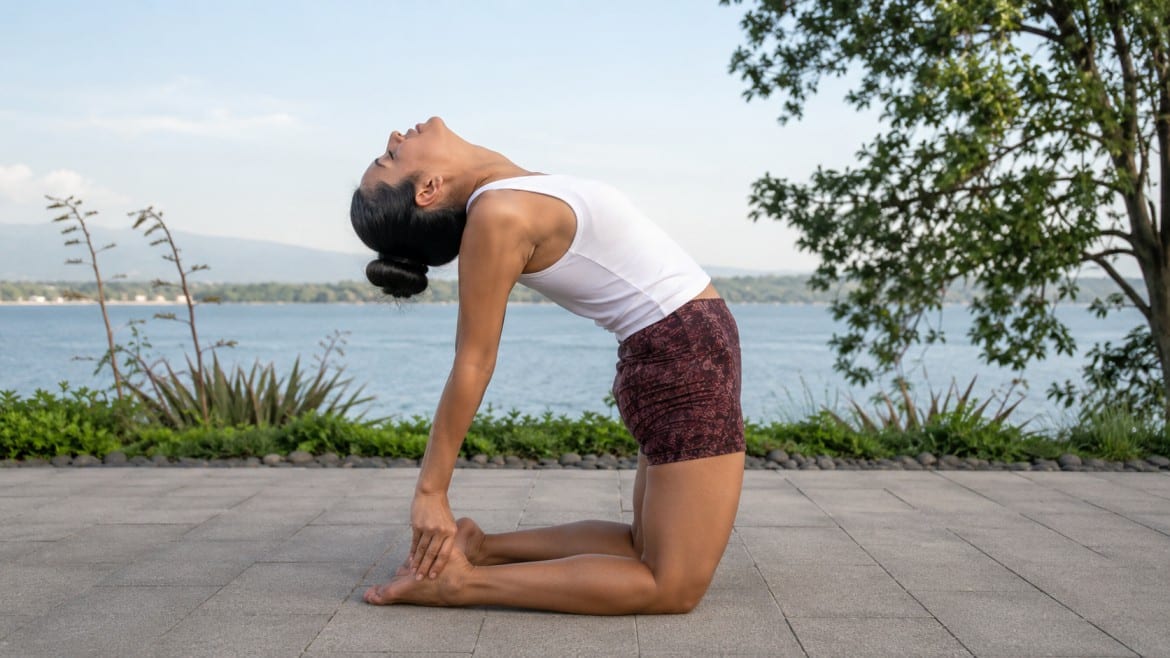 a woman performs a camel pose yoga stretch on a paved surface by a tranquil lake