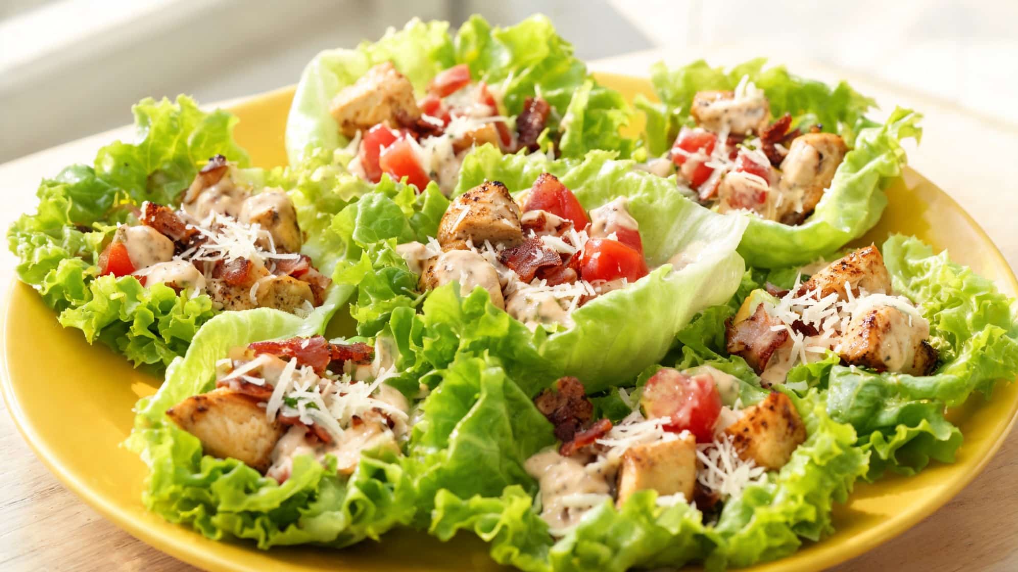 a yellow plate features fresh lettuce cups filled with grilled chicken, diced tomatoes, bacon bits, and grated cheese, creating a vibrant, healthy dish