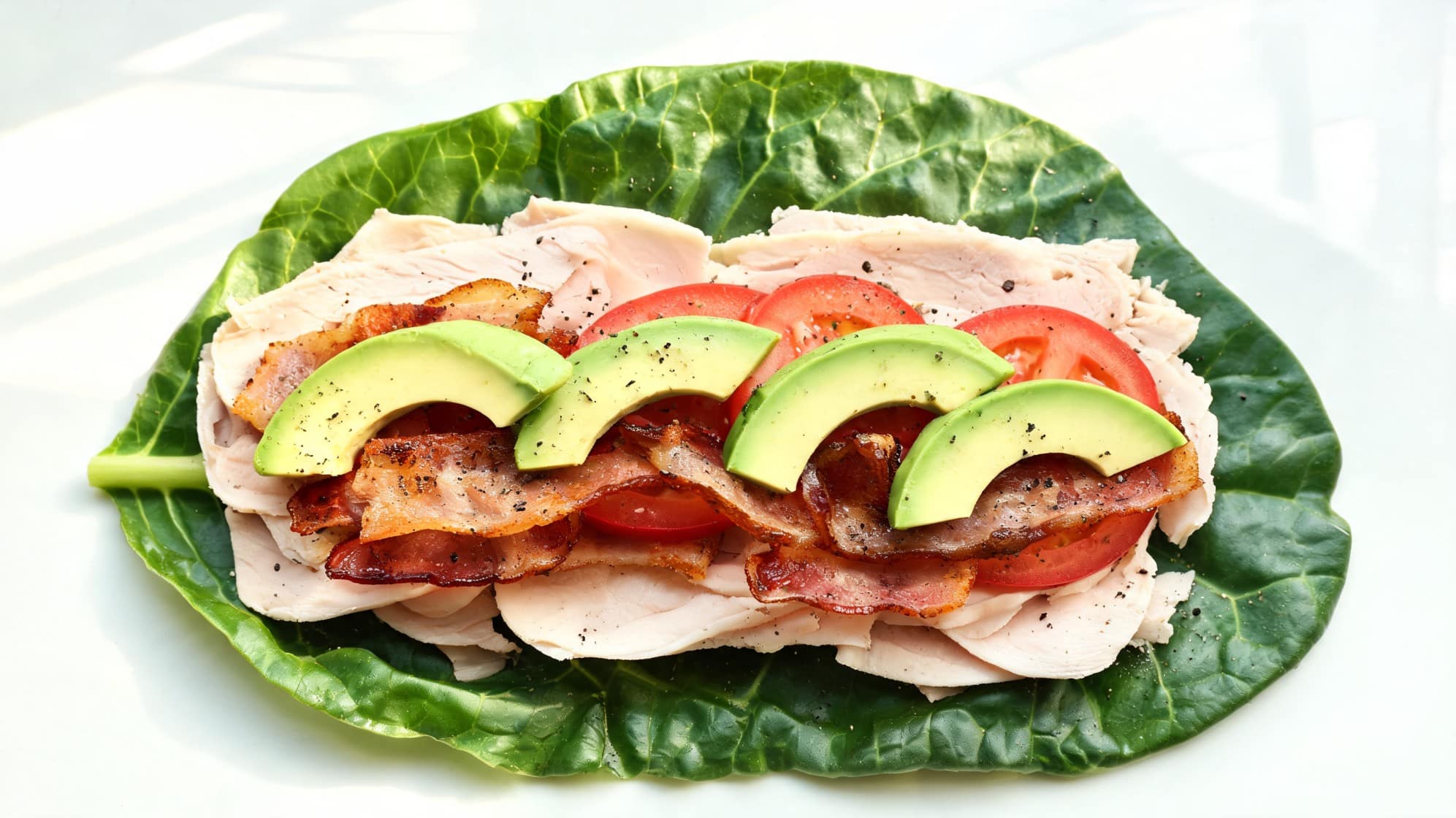 an open-faced sandwich with turkey slices, collard, bacon, and avocado on a large leaf