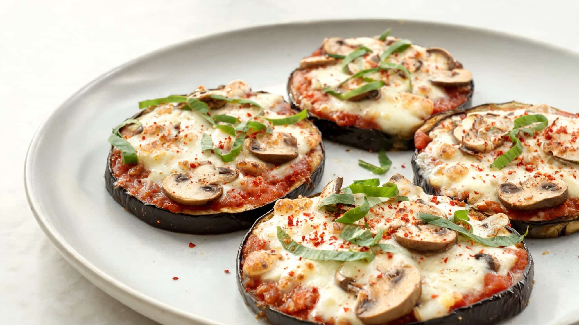 Close-up of a white plate with four eggplant slices topped with melted cheese, tomato sauce, mushrooms, and fresh basil (1)
