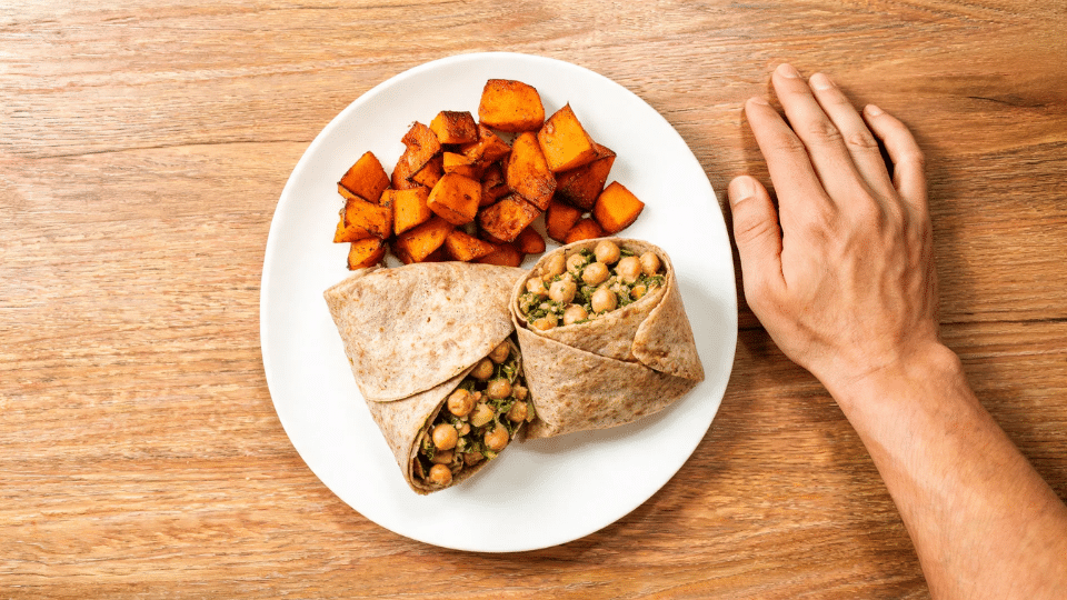 hand beside a plate with roasted sweet potatoes and veggie wrap on wooden table