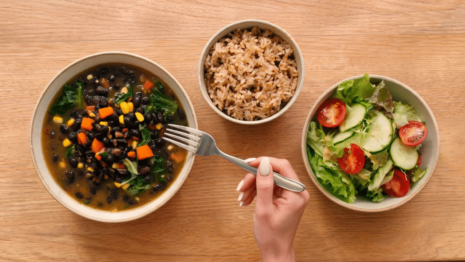 hand holding a fork over bowls of beans rice and salad on a wooden table