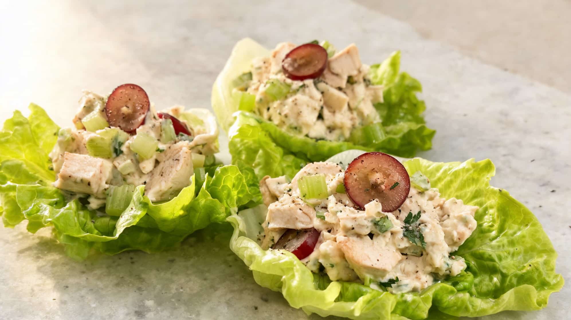 Lettuce wraps filled with creamy chicken salad, topped with sliced grapes and celery on a stone surface. (1)