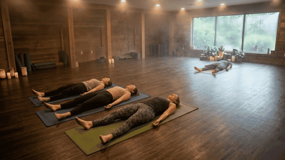 people lying in corpse pose on mats in a dimly lit steamy hot yoga studio with wood paneling