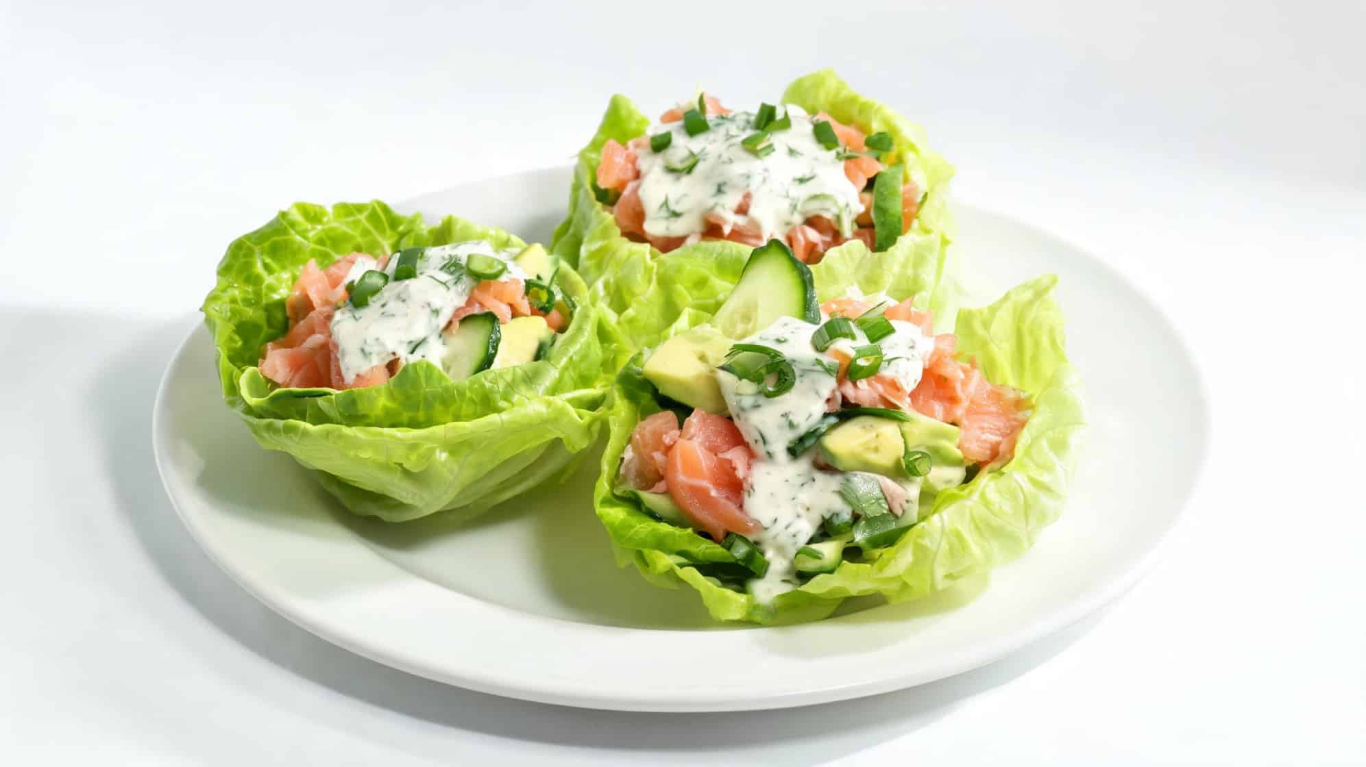 Three lettuce cups filled with fresh salmon, avocado, and cucumber slices, topped with creamy dressing and herbs