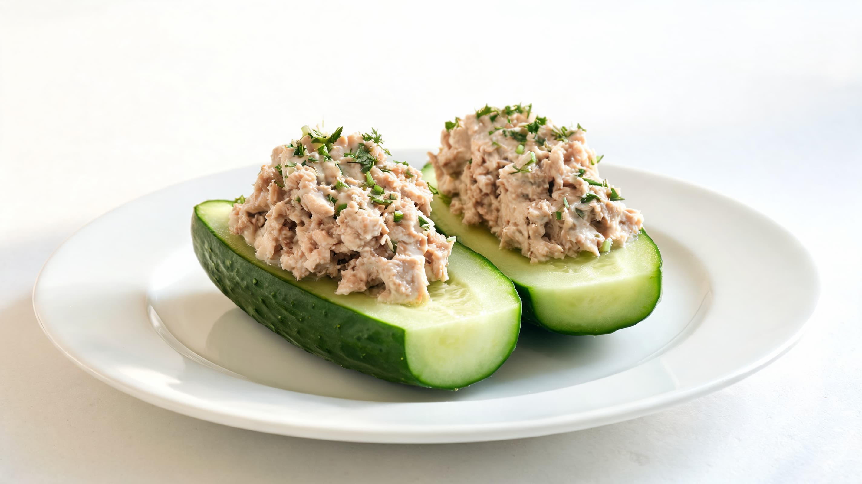 Two cucumber halves on a white plate, filled with a creamy tuna salad. Garnished with herbs