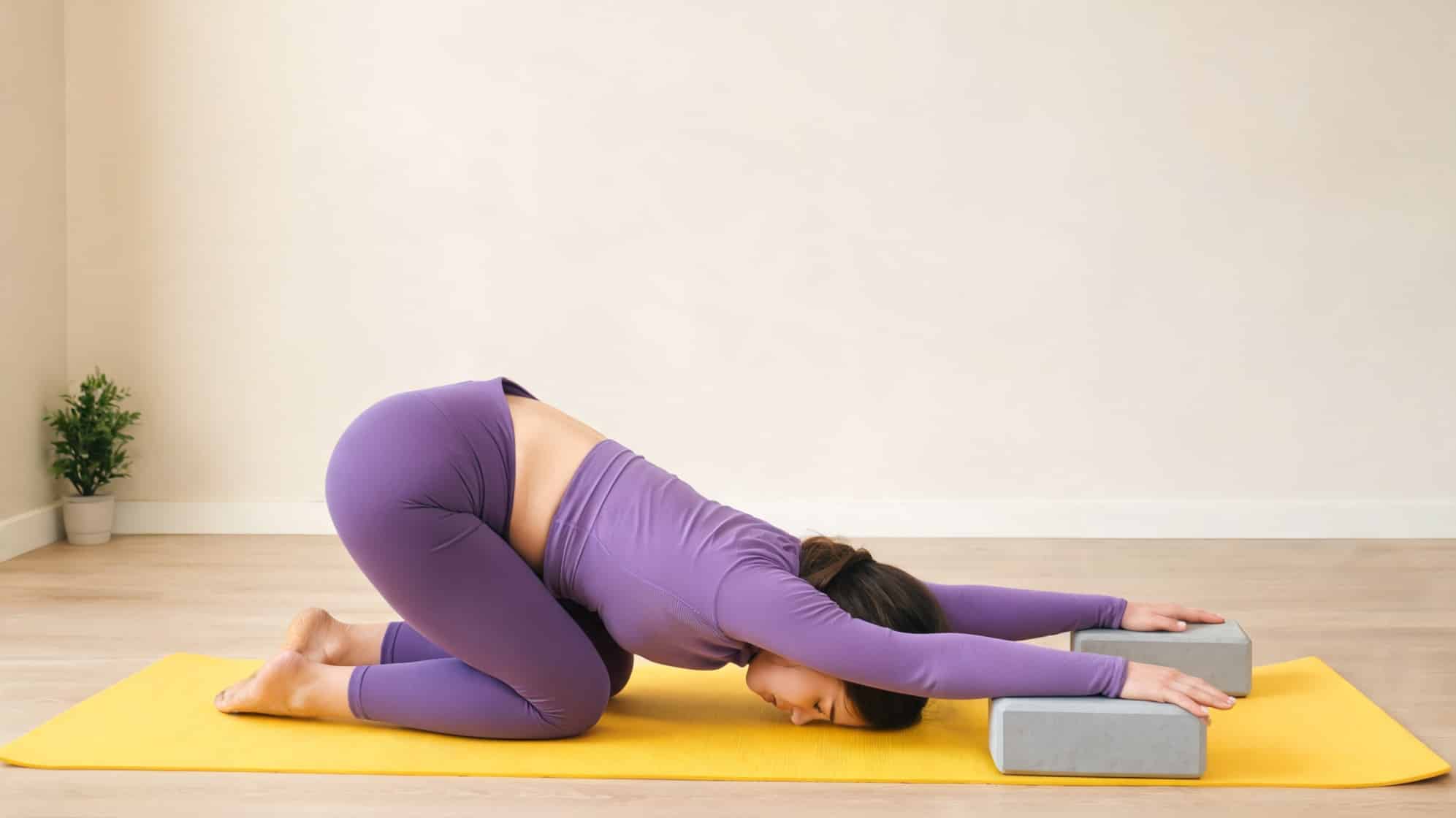 woman in a purple workout outfit performs a child’s pose on a yellow yoga mat, her hands resting on yoga blocks
