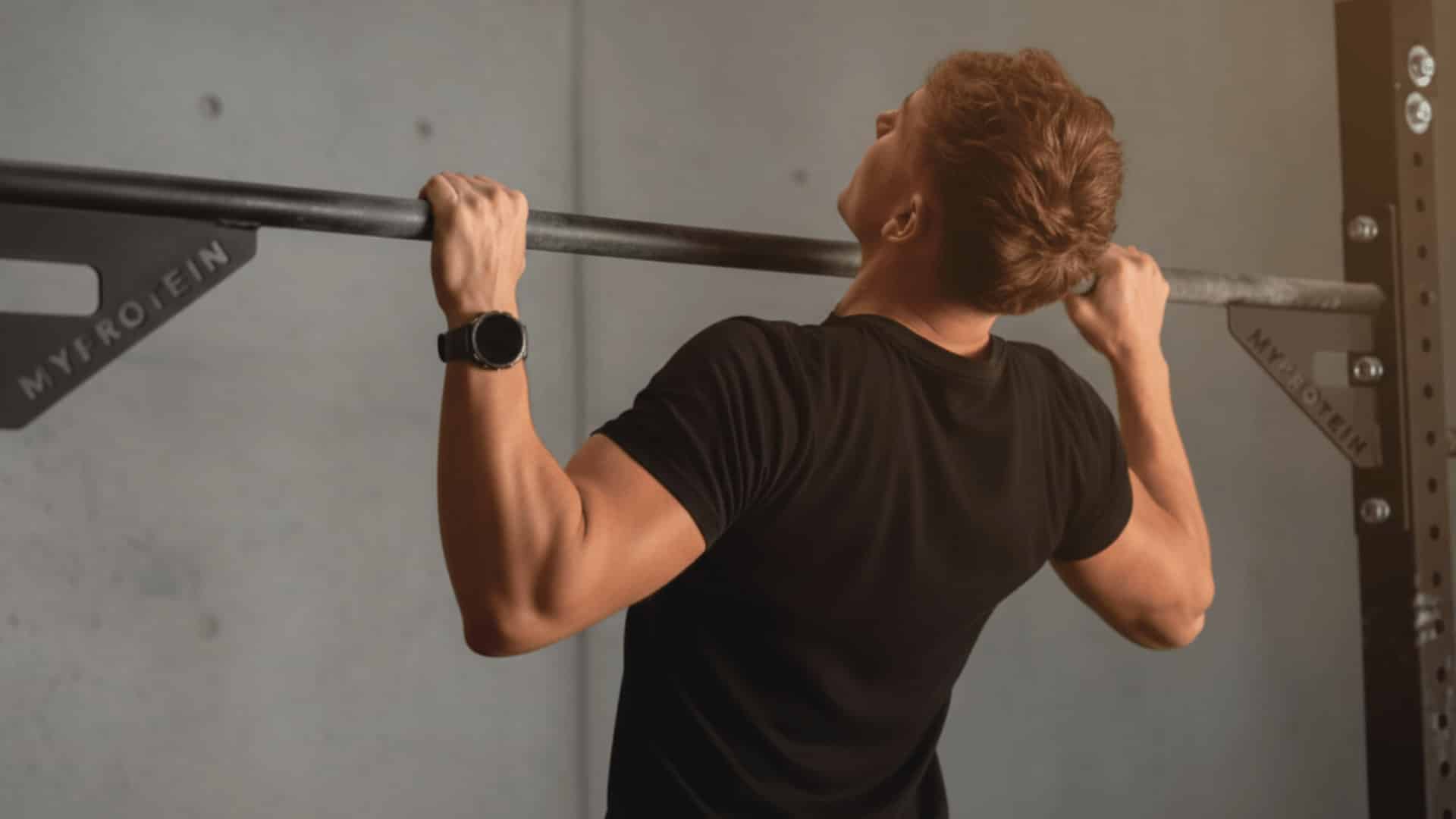 a man performing a pull up grip variation on a barbell rack in a gym, back view