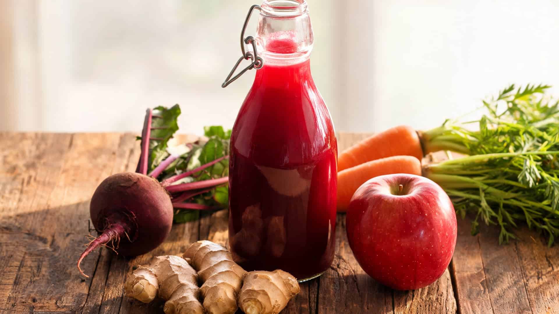 fresh beet juice in a glass bottle surrounded by ginger carrot apple and leafy greens on wooden surface