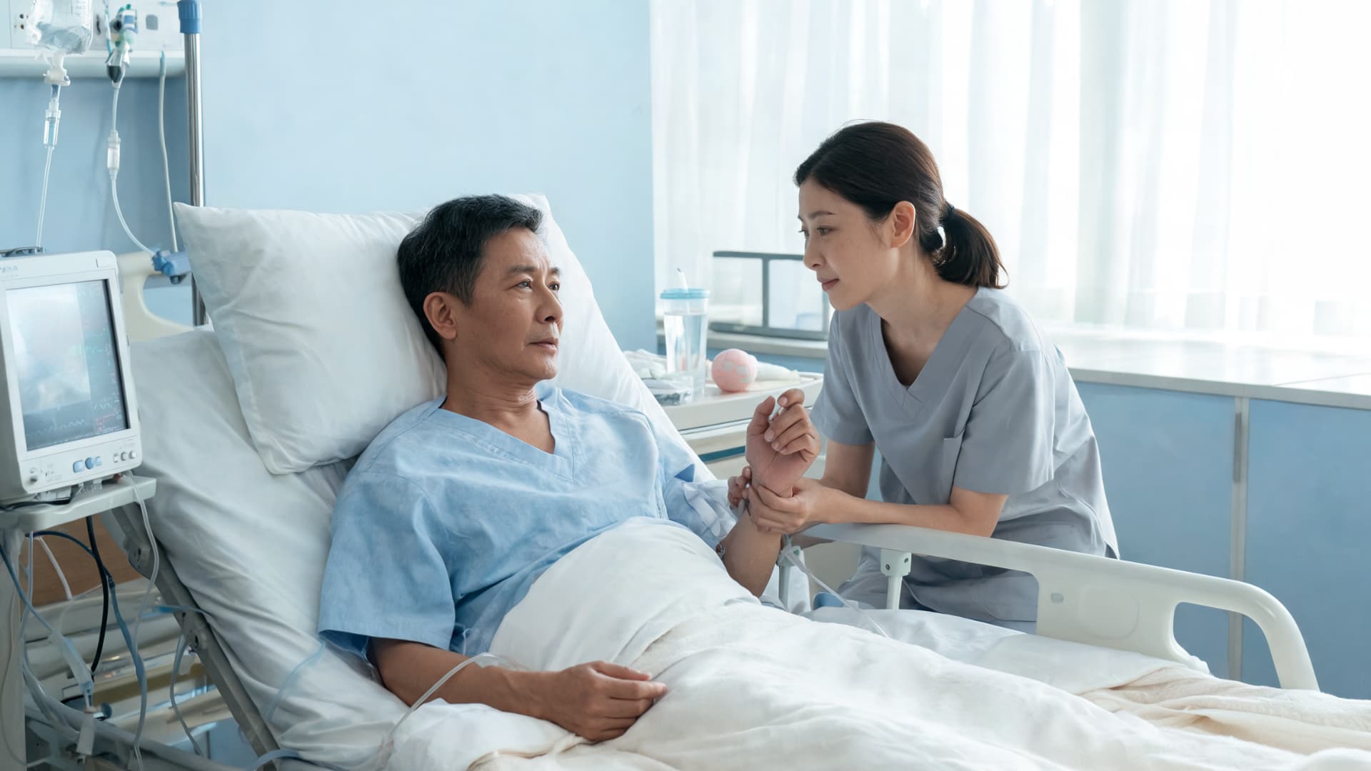 male patient lying in hospital bed with monitoring while a female nurse holds his hand