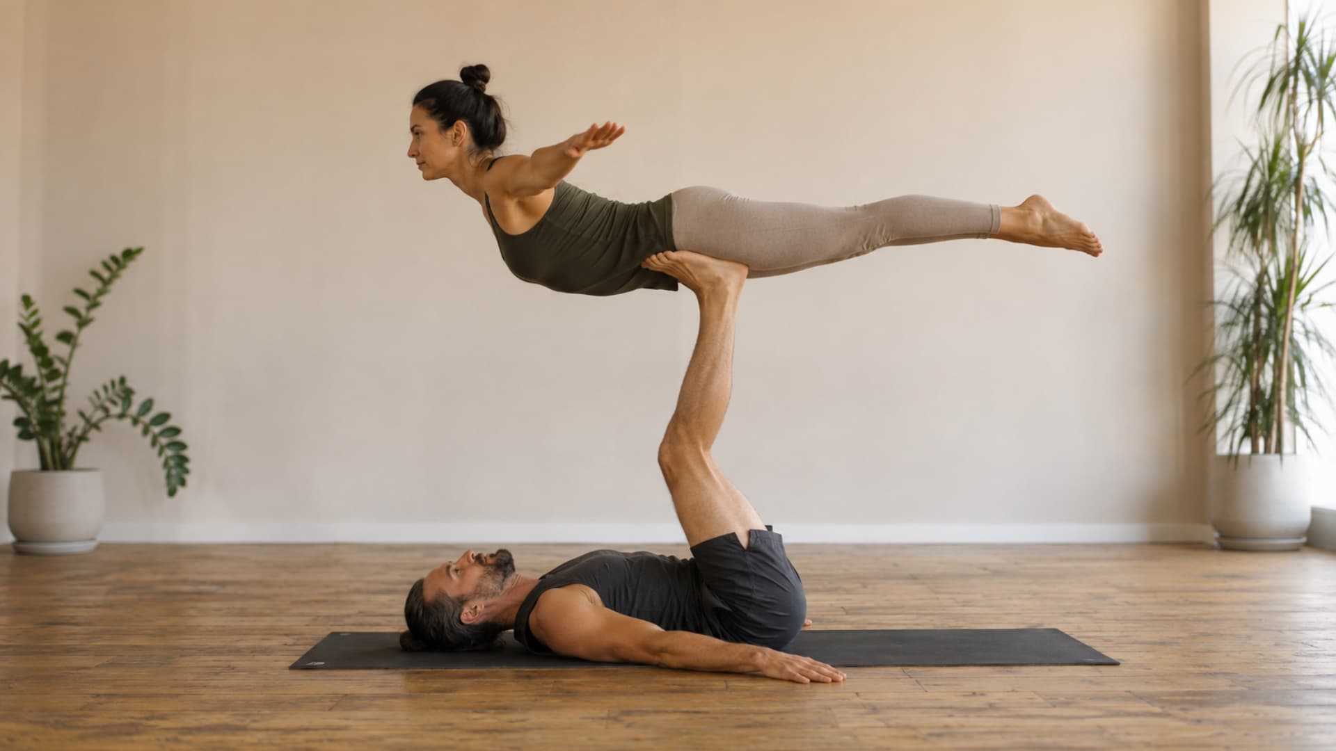 Two adults practice bird pose in a calm yoga studio with strong balance and steady acro support