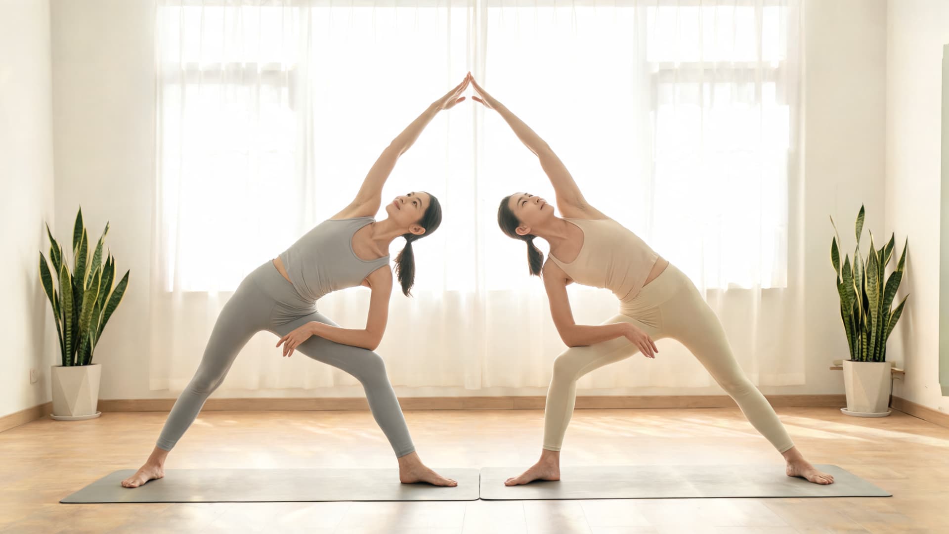 Two partners hold extended side angle pose with raised hands touching in a bright peaceful yoga studio