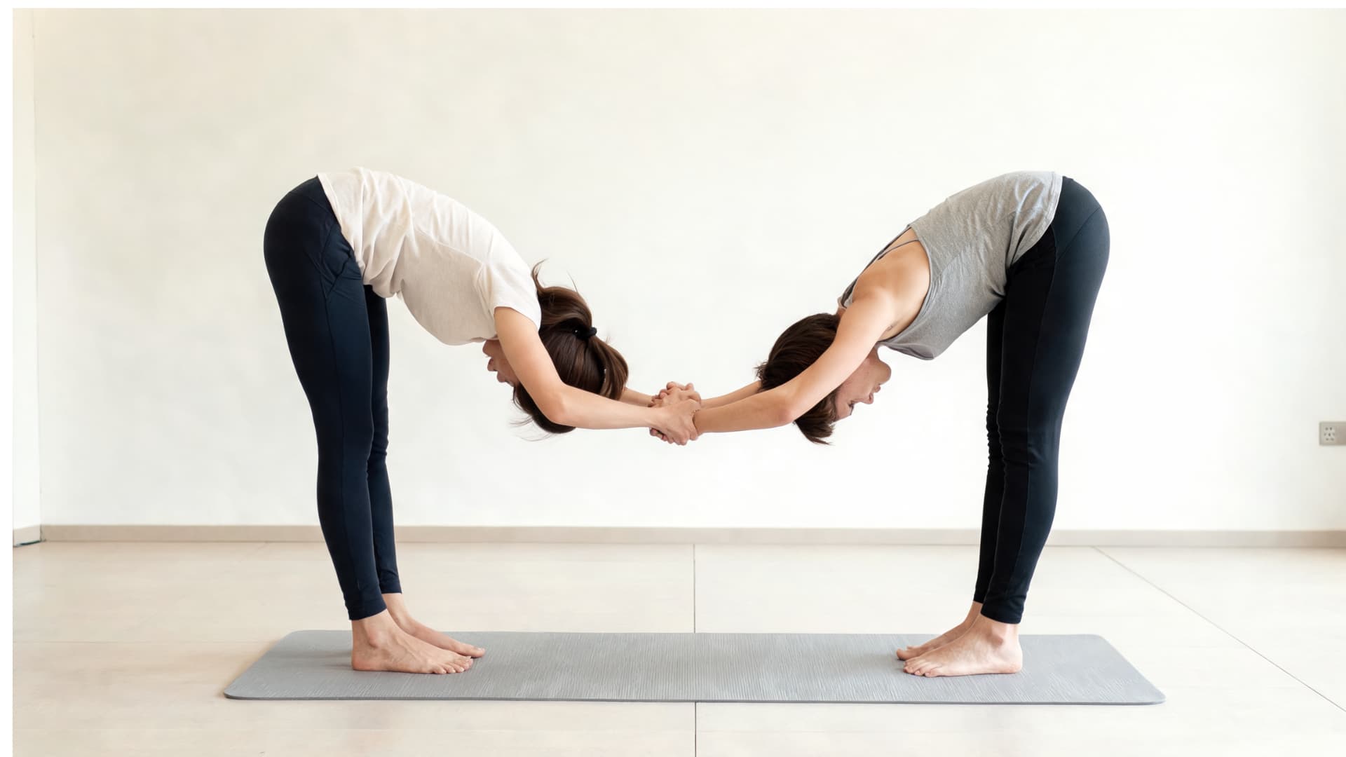 Two people practicing standing partner forward fold with hands joined on a yoga mat in a bright studio