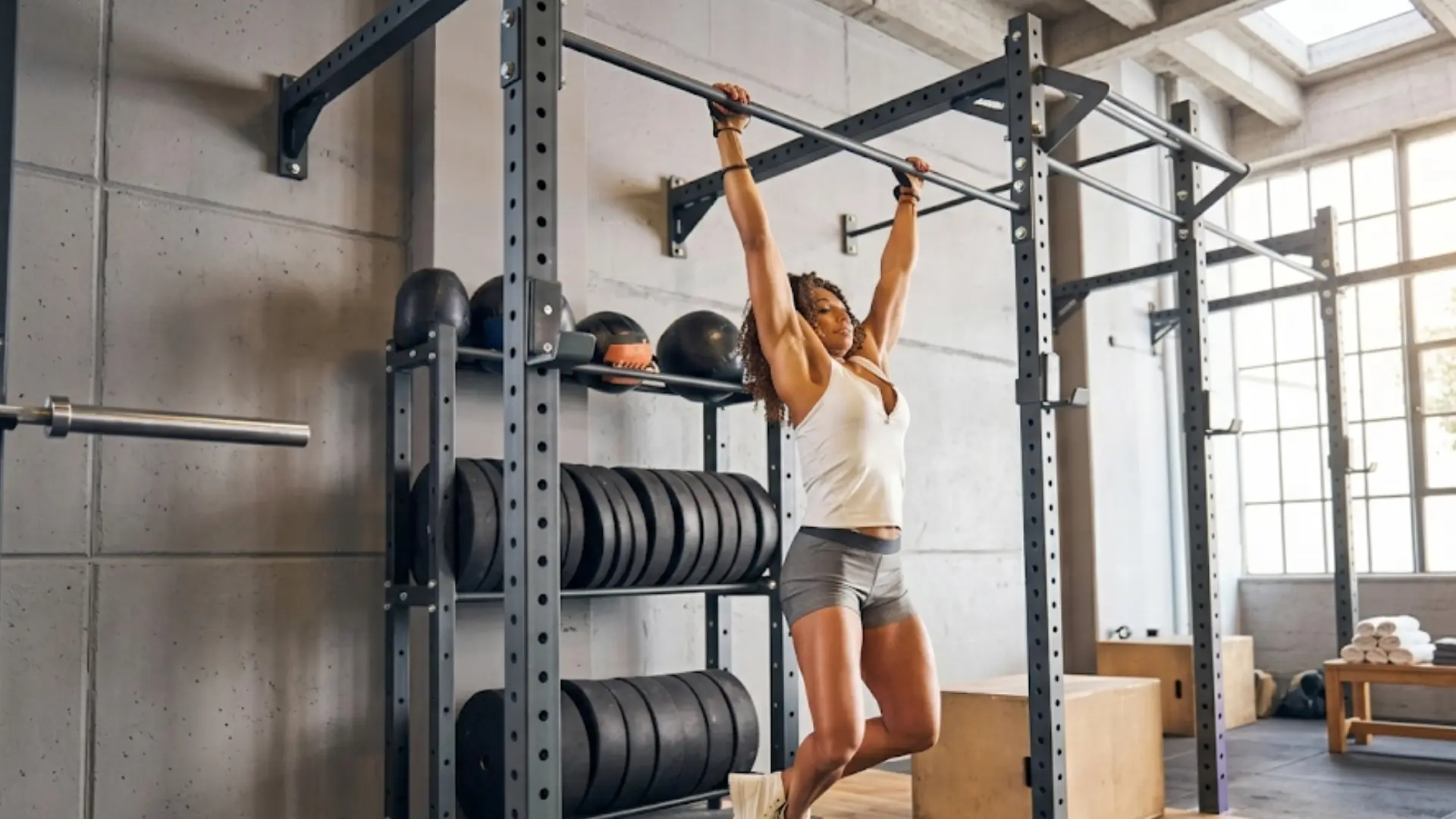 woman hangs from a pull-up bar in a spacious, sunlit gym filled with weight plates and workout rigs