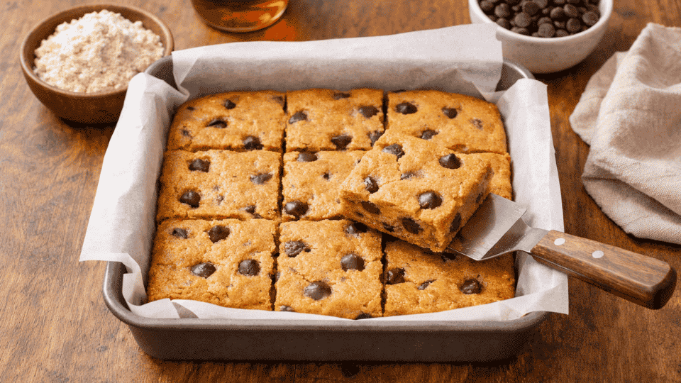 an of paleo chocolate chip blondies cut into squares, one piece lifted on spatula, with almond flour and chocolate chips nearby